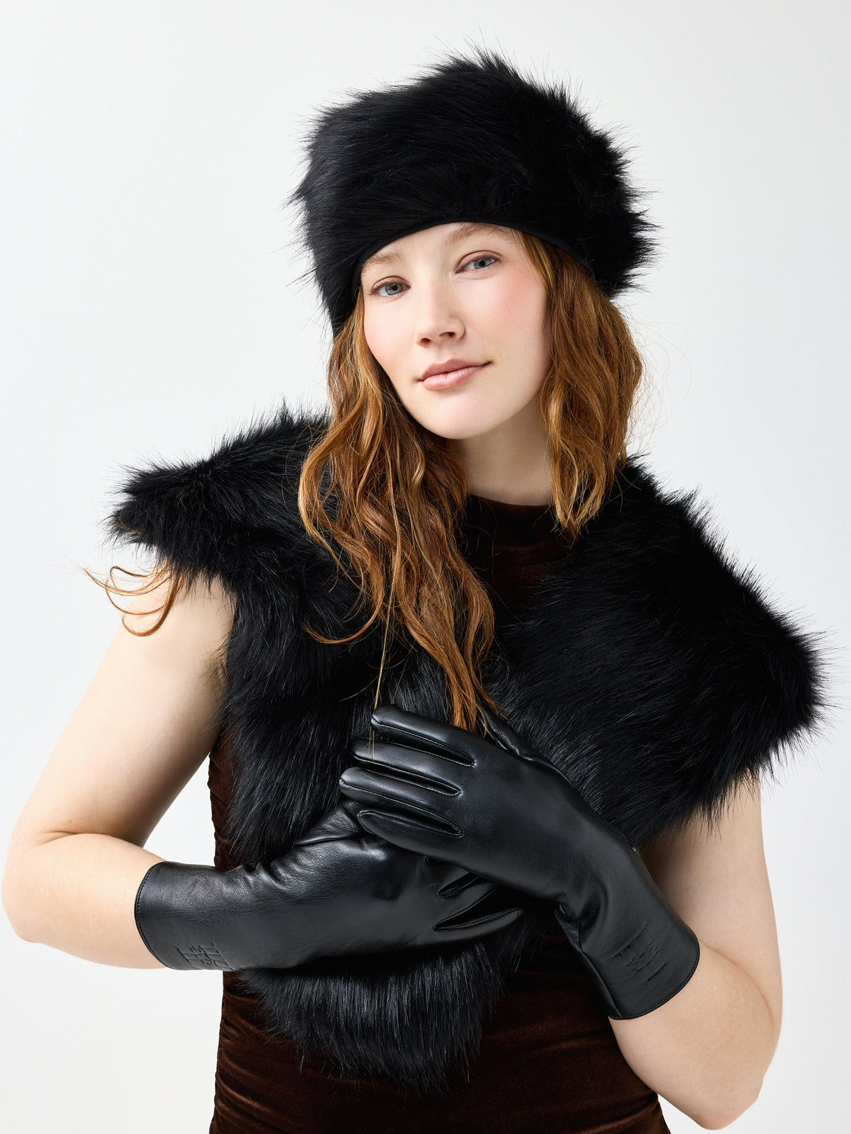 Woman wearing a black faux fur hat, black vegan leather gloves, and a black faux fur scarf on a white background