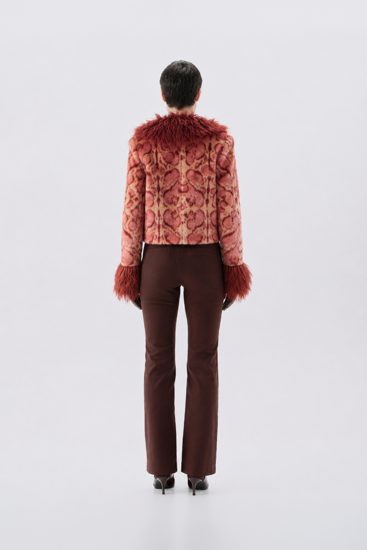 A model shown from the back wearing a cropped, patterned pink jacket with reddish-brown faux fur trim at the collar and cuffs, paired with dark brown trousers and black heels, against a light gray studio background.
