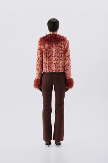A model shown from the back wearing a cropped, patterned pink jacket with reddish-brown faux fur trim at the collar and cuffs, paired with dark brown trousers and black heels, against a light gray studio background.