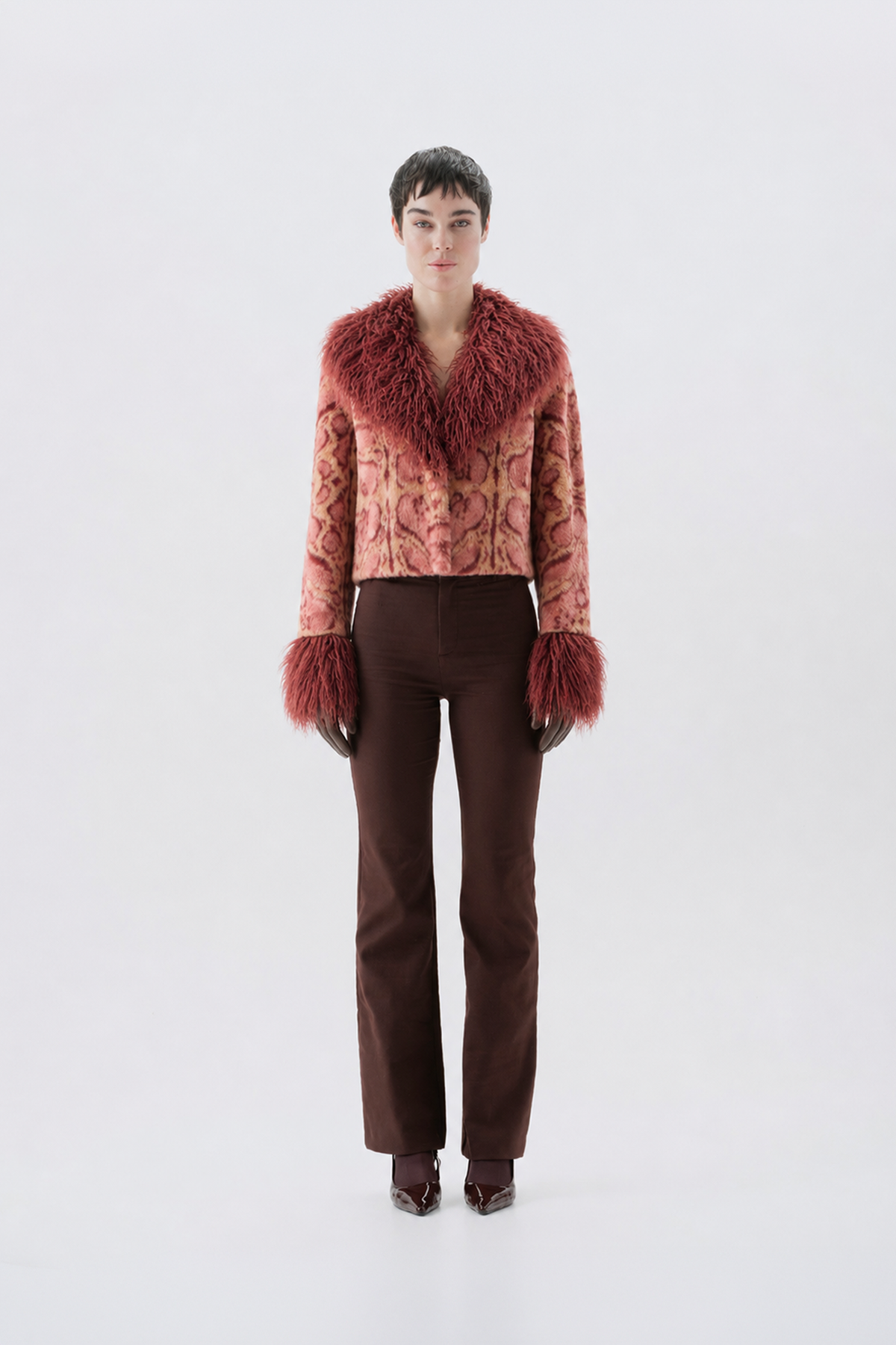 A model wearing a cropped pink patterned jacket with a large reddish-brown fur collar and matching fur-trimmed cuffs, paired with dark brown pants and dark shoes.