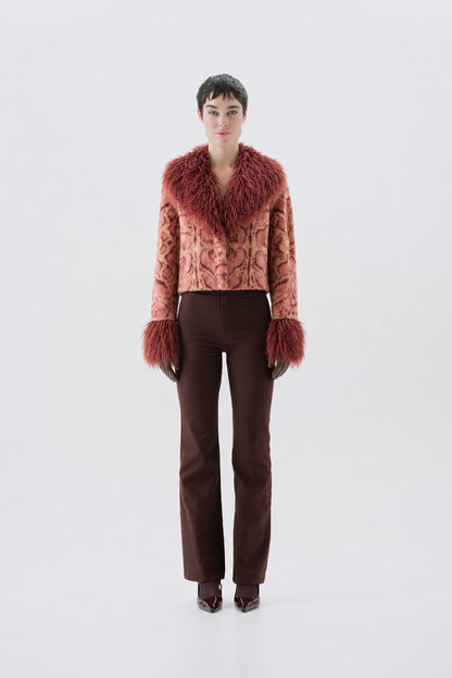 A model wearing a cropped pink patterned jacket with a large reddish-brown fur collar and matching fur-trimmed cuffs, paired with dark brown pants and dark shoes.