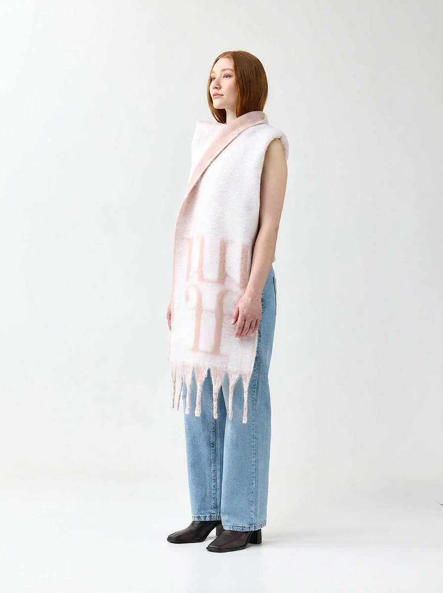 Woman wearing a light pink and white patterned scarf with fringe, standing against a white background.